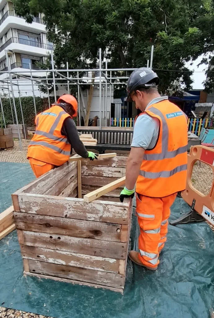 Two workers in high-visibility vests assembling a planter box from salvaged wood for the Ebury Bridge Estate community garden